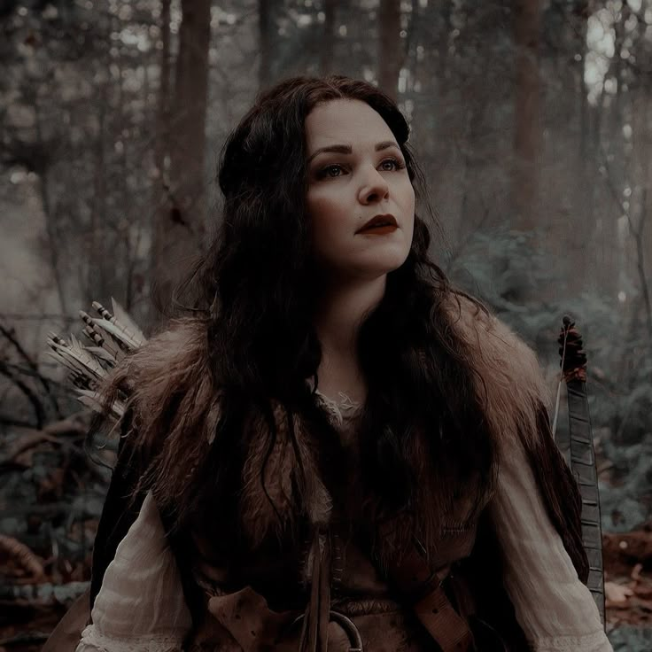 a woman with long hair wearing a fur vest and holding a bow in the woods