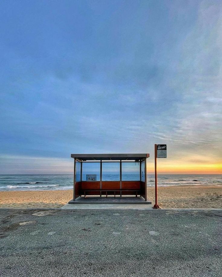 a bus stop sitting on top of a sandy beach next to the ocean at sunset