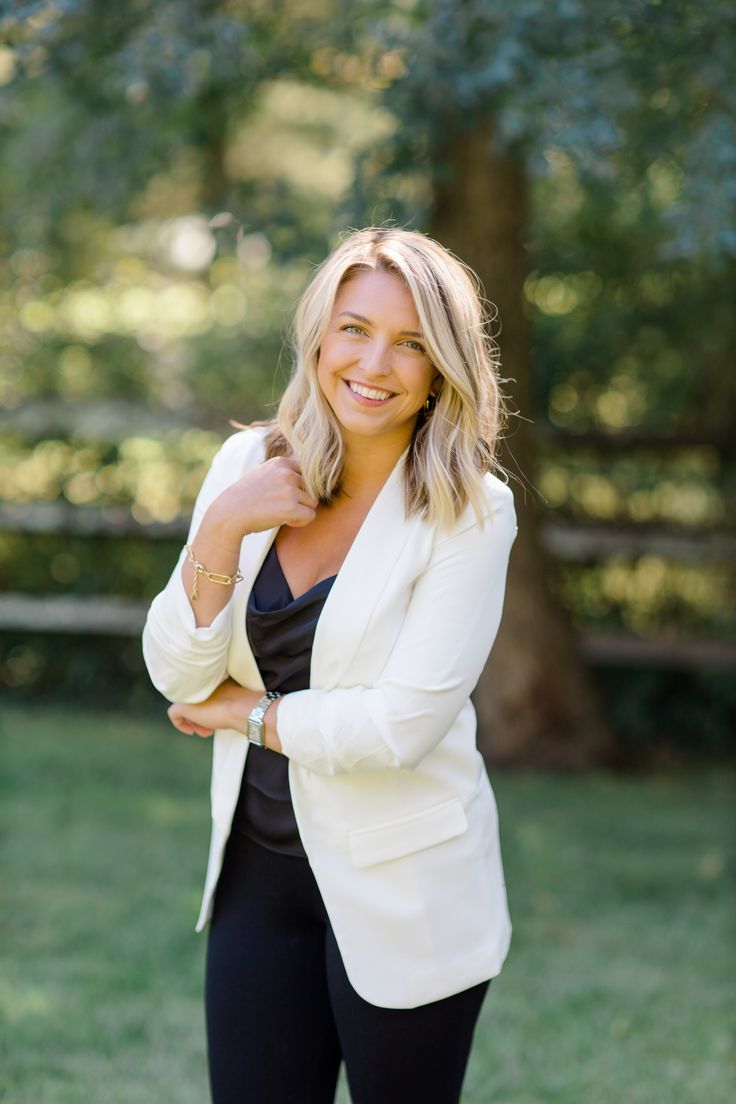 a woman in black pants and a white blazer smiles at the camera with her arms crossed