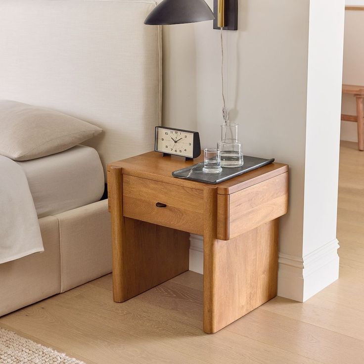 a small wooden table with a clock on it next to a bed and nightstand in a bedroom