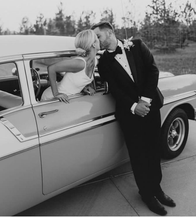 a man in a tuxedo leaning on the back of a car with a woman kissing him