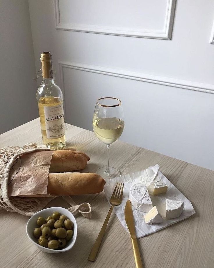 a table with bread, cheese and olives next to a glass of white wine