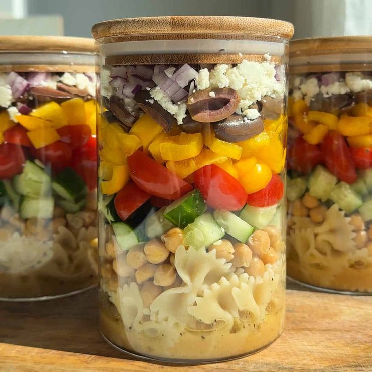 three mason jars filled with different types of vegetables and pasta salad in them on a wooden table