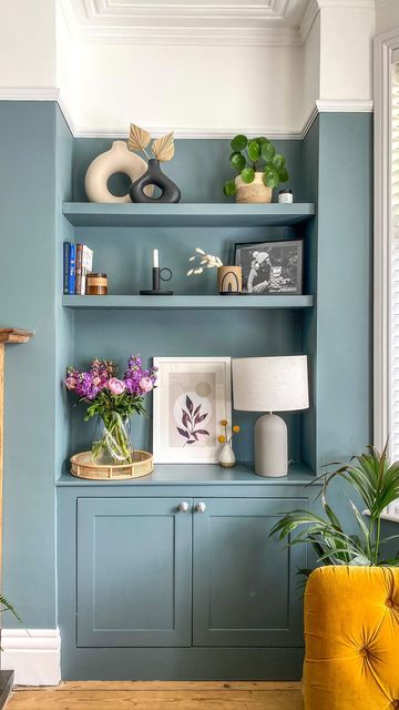 a living room with blue painted walls and shelves filled with flowers, plants and pictures