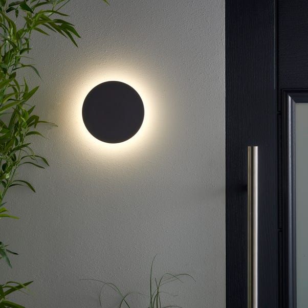 a round light on the side of a wall next to a potted plant and door