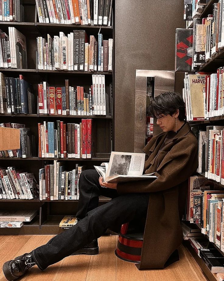 a man sitting on a stool in front of a bookshelf reading a book