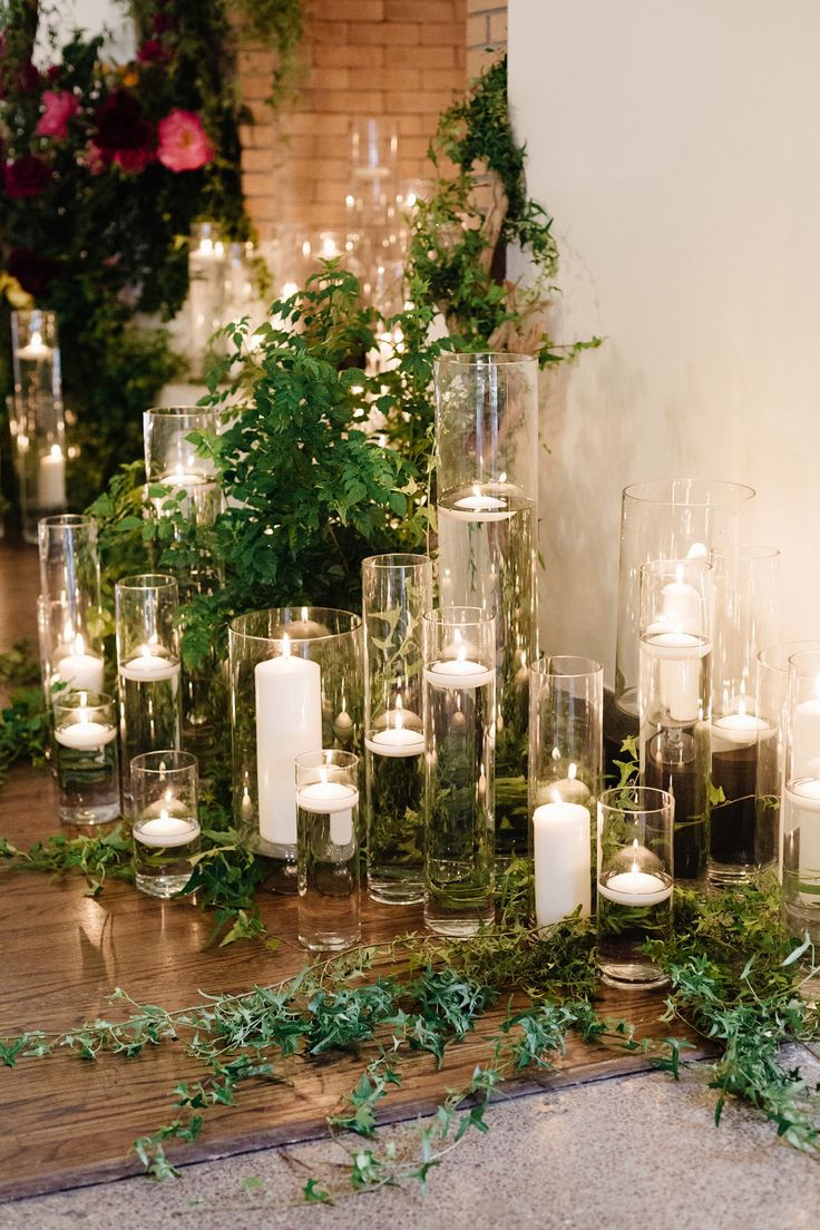 candles and greenery are arranged in glass vases on the floor next to a brick wall