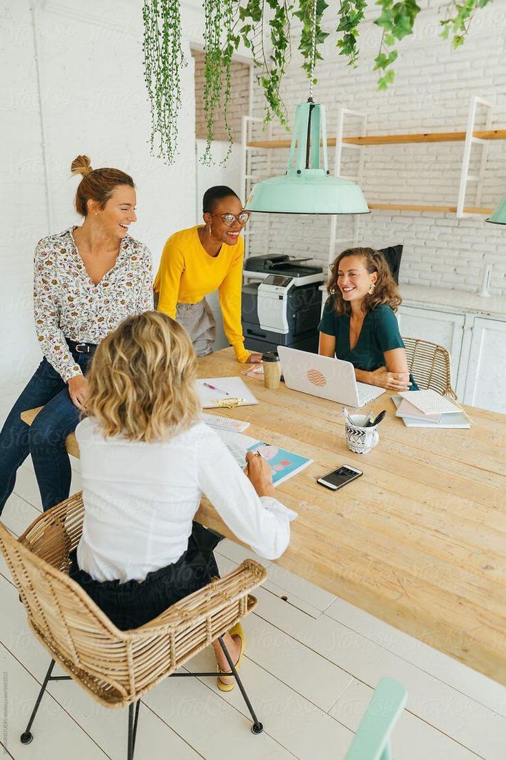 three women sitting at a table with laptops and papers in front of them by an office