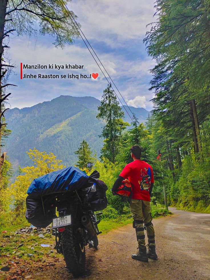 a man standing next to a motorcycle on a dirt road in the woods with mountains behind him