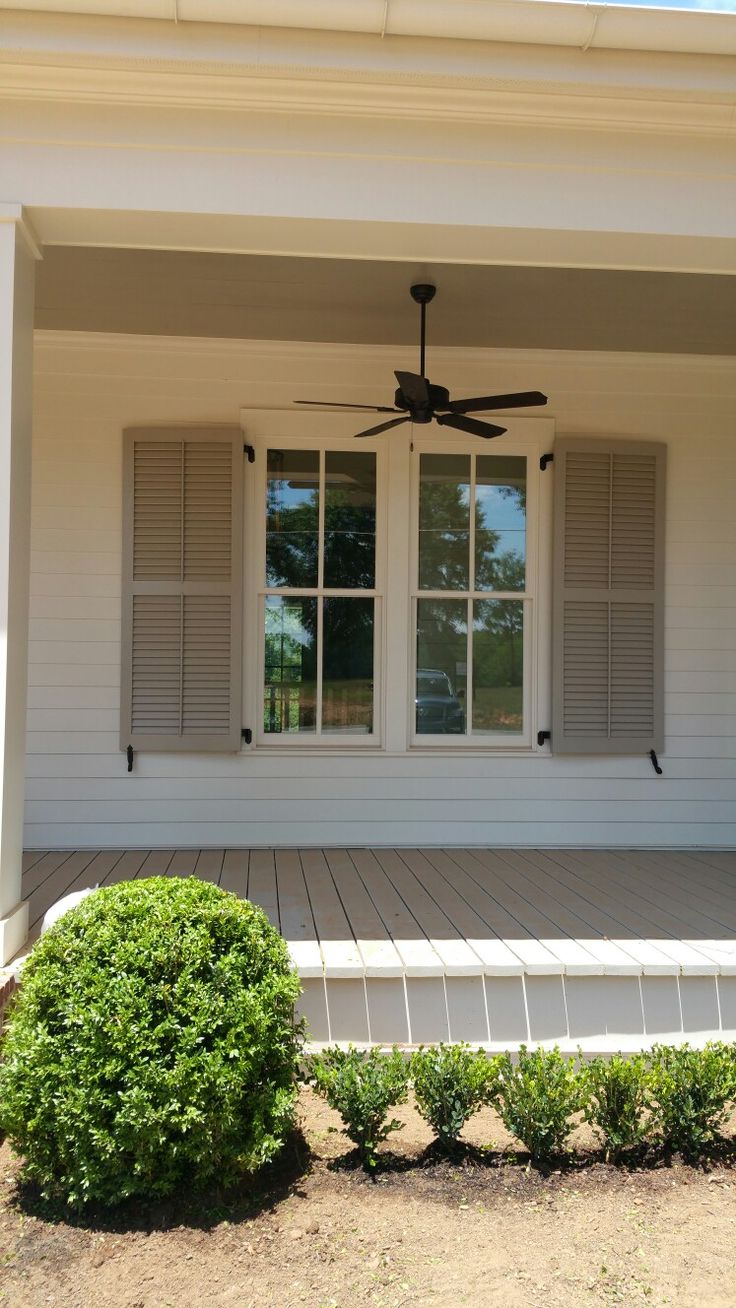 a white house with shutters and a ceiling fan on the front porch, surrounded by shrubbery