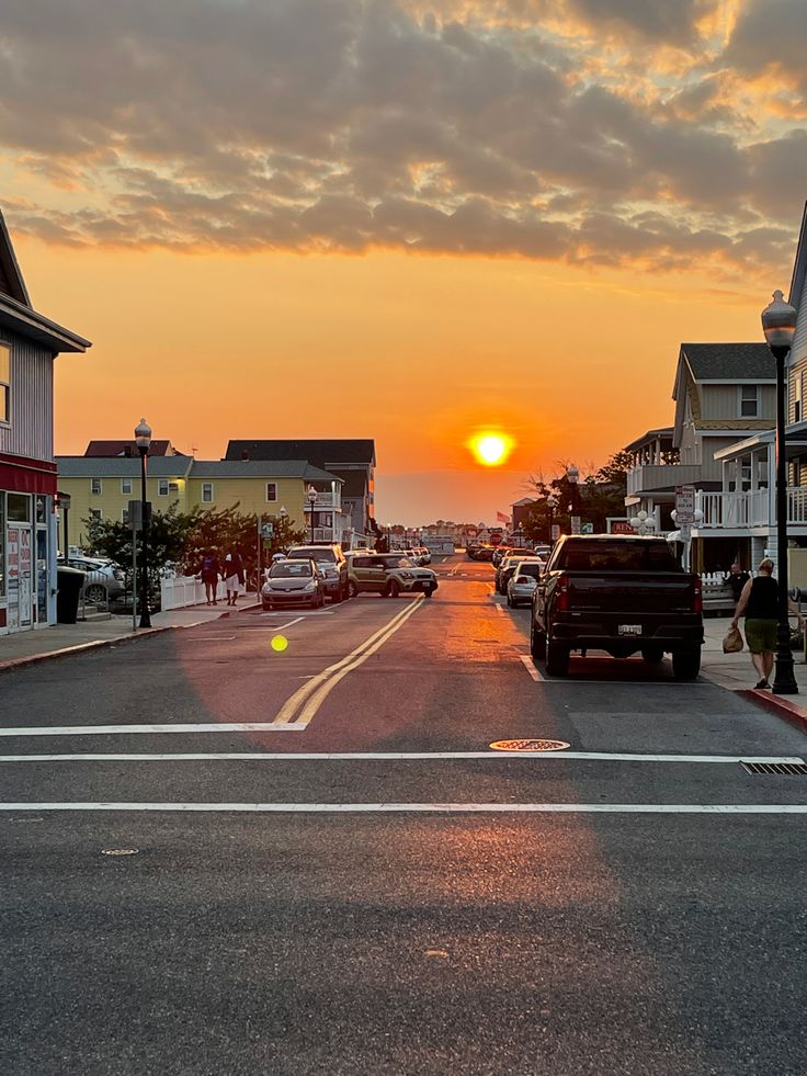 the sun is setting on an empty street with cars parked in front of houses and buildings