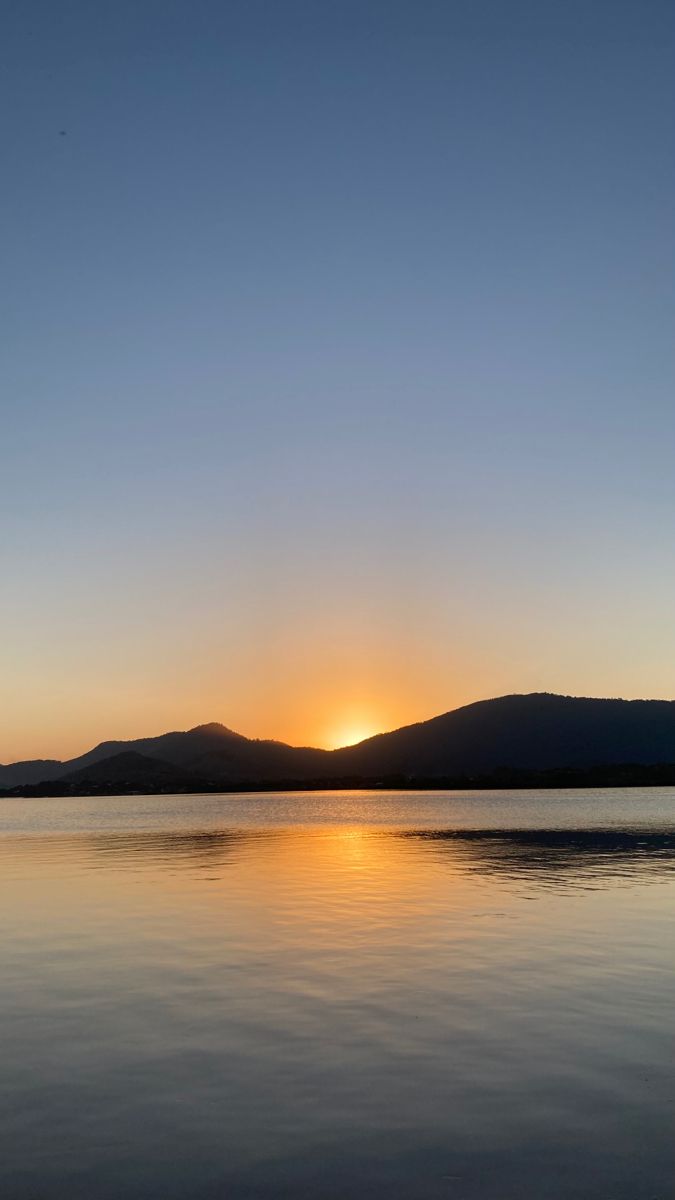 the sun is setting over water with mountains in the distance and blue sky above it