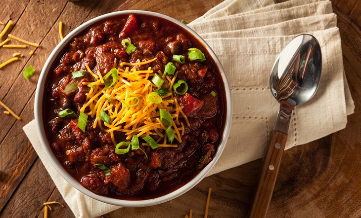 a bowl of chili with cheese and green onions next to a spoon on a napkin