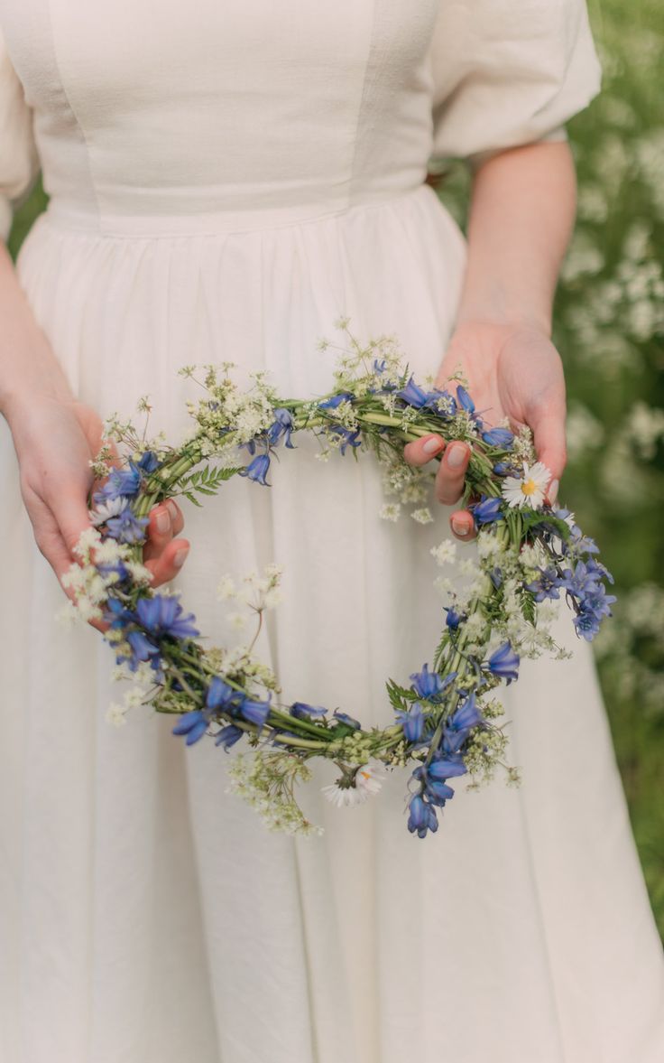 a woman in a white dress holding a blue and white flower wreath with her hands