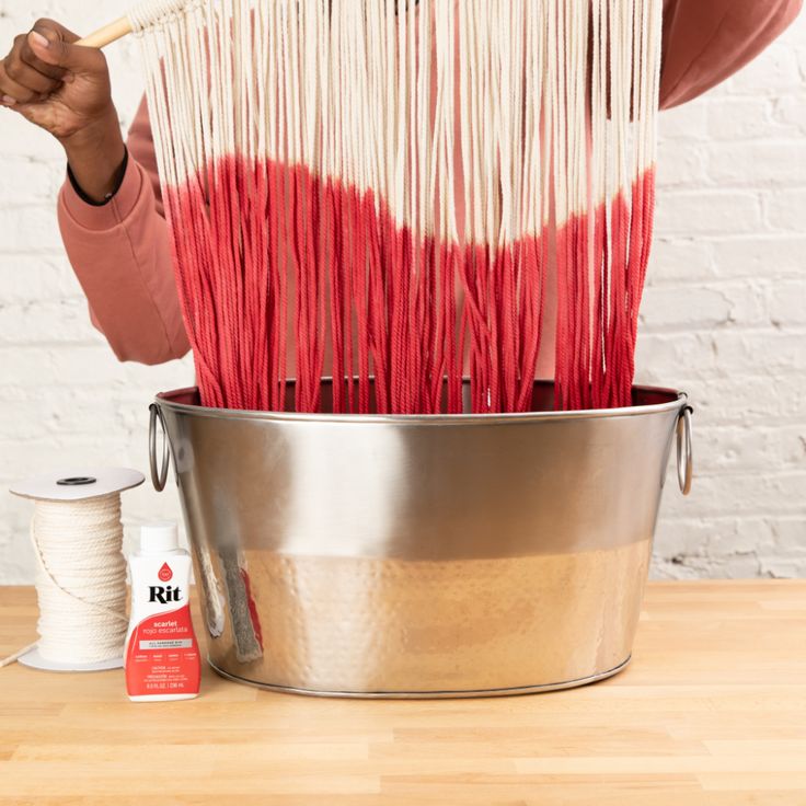 a woman is weaving red and white yarn in a metal bucket on a wooden table