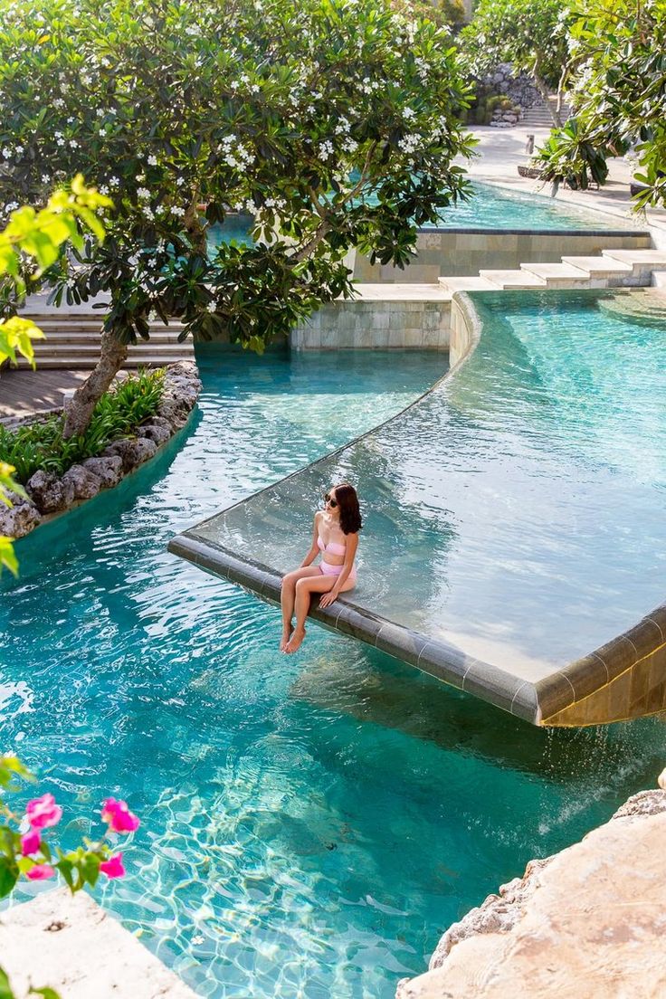 a woman sitting on the edge of a swimming pool next to trees and steps in front of her