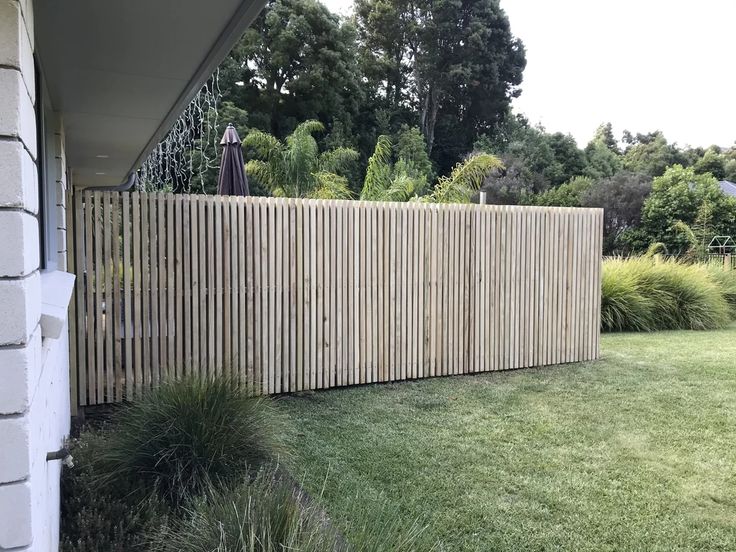 a wooden fence in front of a house with grass on the lawn and bushes behind it