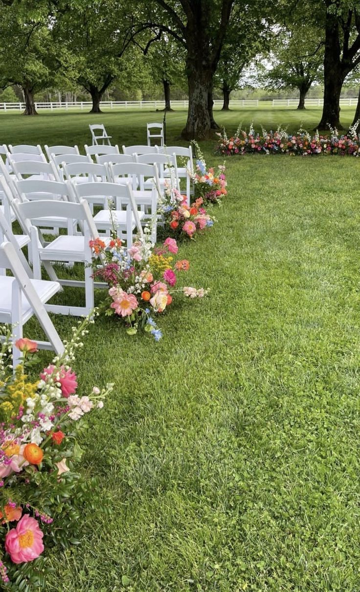 rows of white chairs lined up in the grass with flowers on each row and pink, red, orange, and yellow flowers at the end