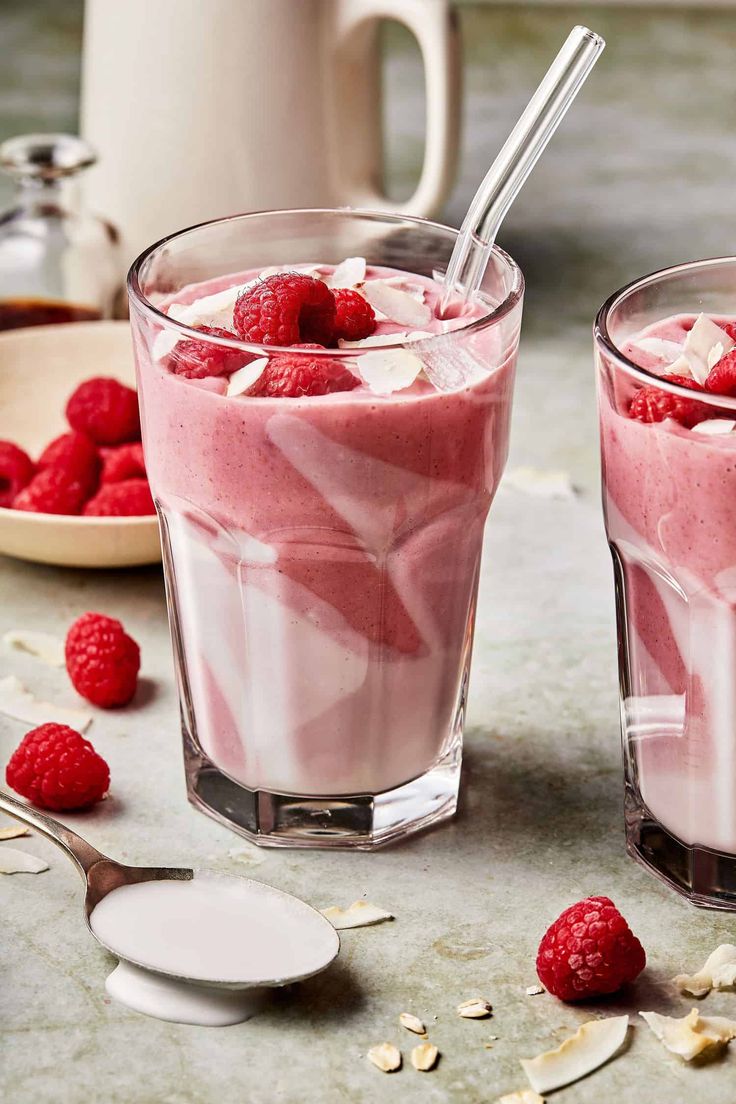 two glasses filled with raspberry smoothie next to some raspberries on the table