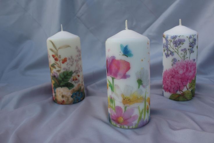 three candles with flowers painted on them are lined up against a blue background, one is white and the other is pink