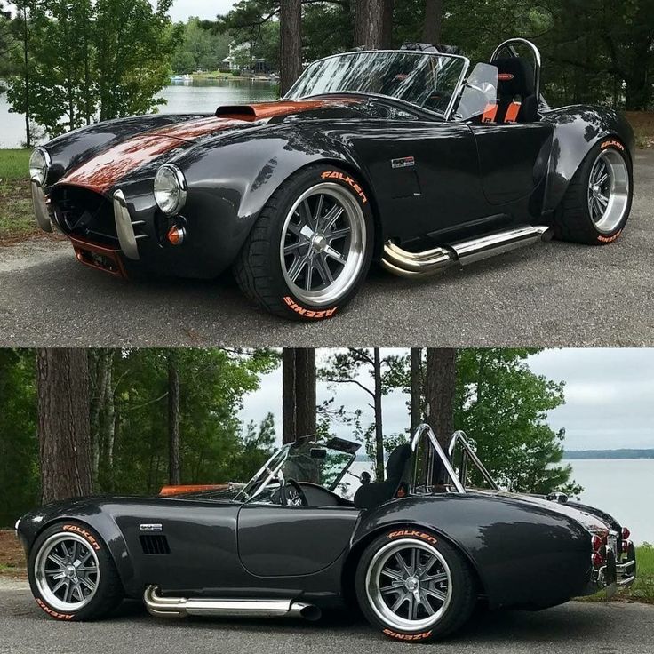 an old black sports car parked in front of a lake with trees and water behind it