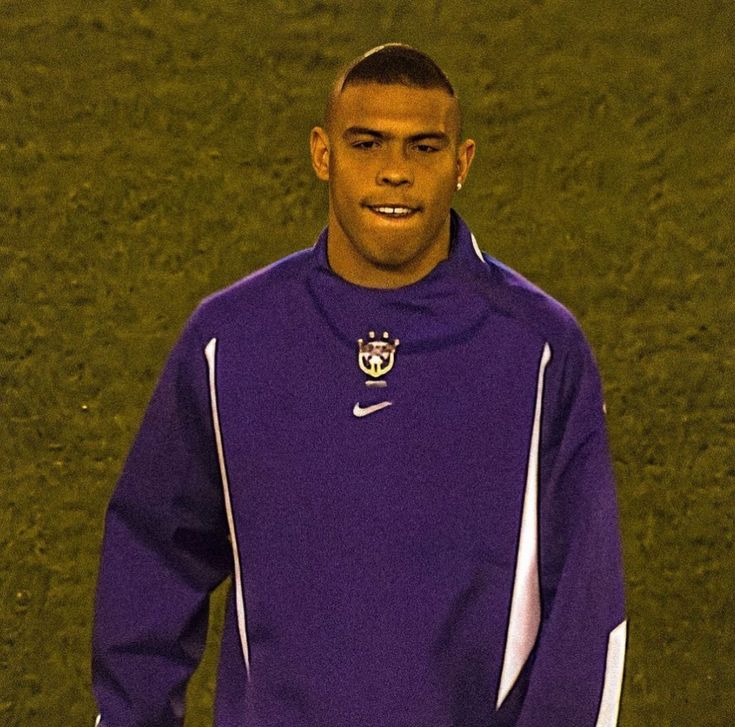 a man in a purple shirt standing on a soccer field with his hands in his pockets