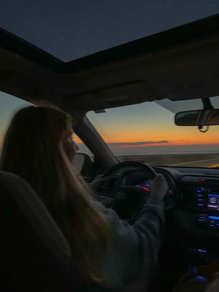 a woman sitting in the driver's seat of a car as the sun sets