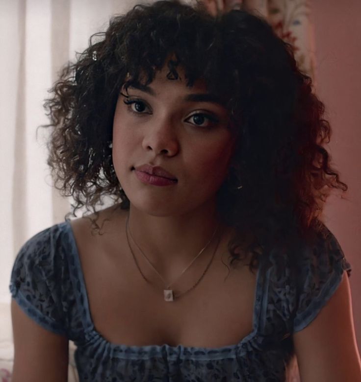 a woman with curly hair wearing a blue dress and looking off to the side while sitting in front of a window