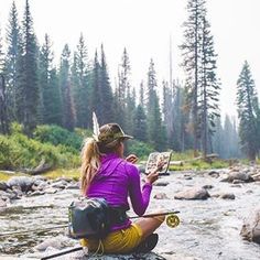 a woman is sitting on rocks in the water and looking at a map while holding a fishing rod