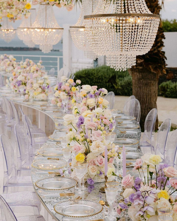 a long table is set up with clear chairs and chandelier hanging from the ceiling