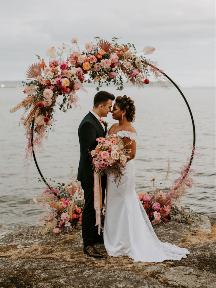 a bride and groom standing in front of an arch with flowers on the water's edge