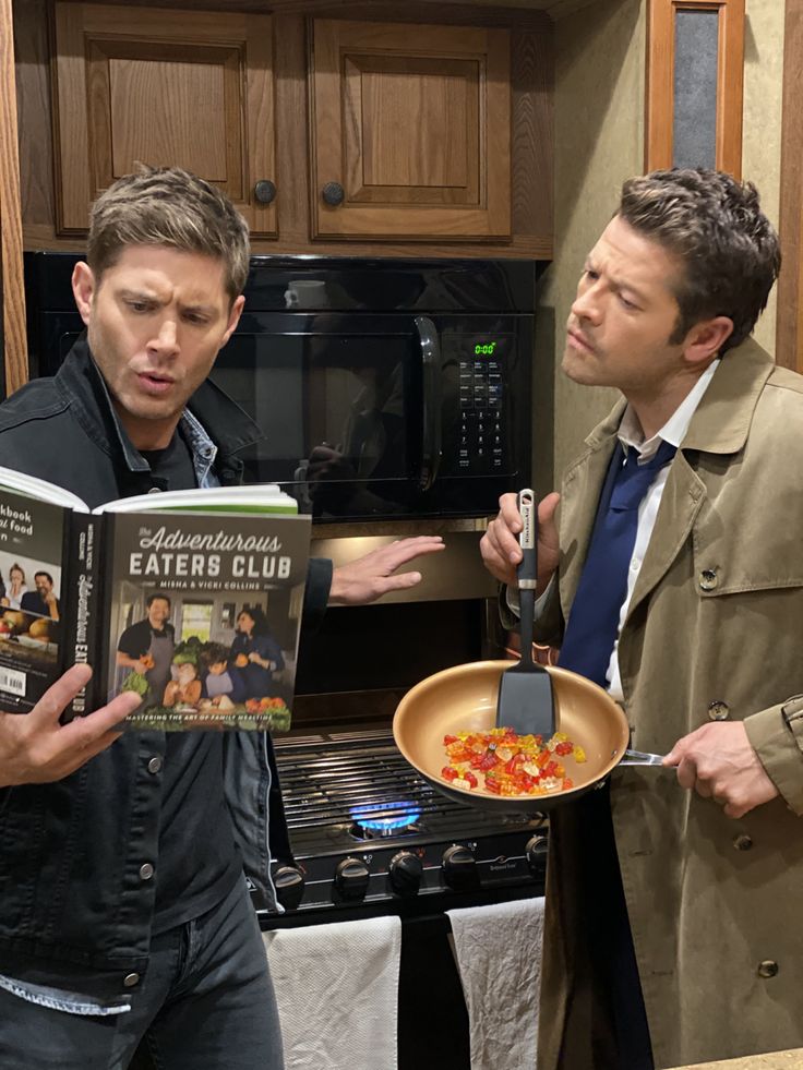 two men standing in a kitchen looking at a cookbook and one holding a frying pan