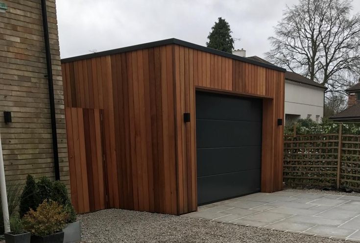 a wooden building with a black door in the middle of a driveway next to a fence