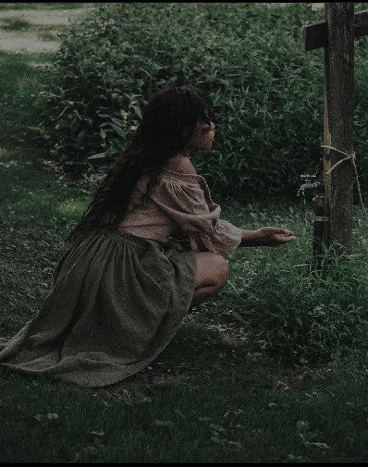 a woman sitting on the ground next to a wooden cross in a green field with grass