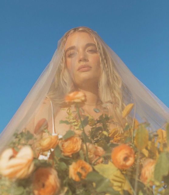 a woman wearing a veil with flowers in front of her face and the sky behind her