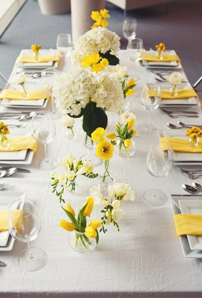 the table is set with yellow and white flowers in vases, napkins, and place settings