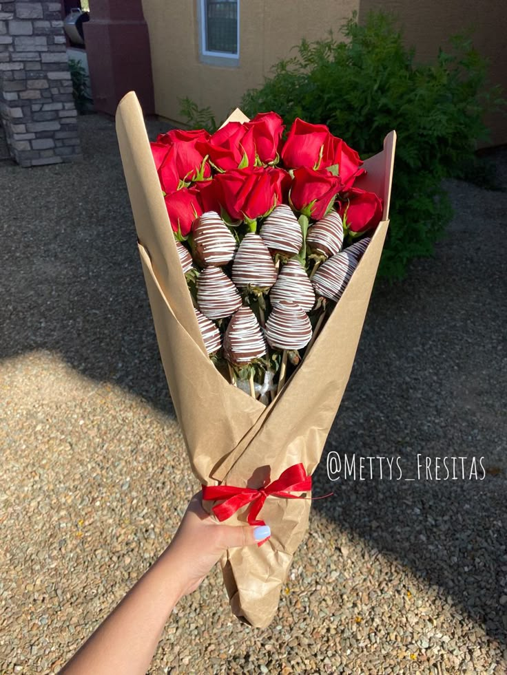 a hand holding a bouquet of roses and chocolate covered strawberries in front of a house