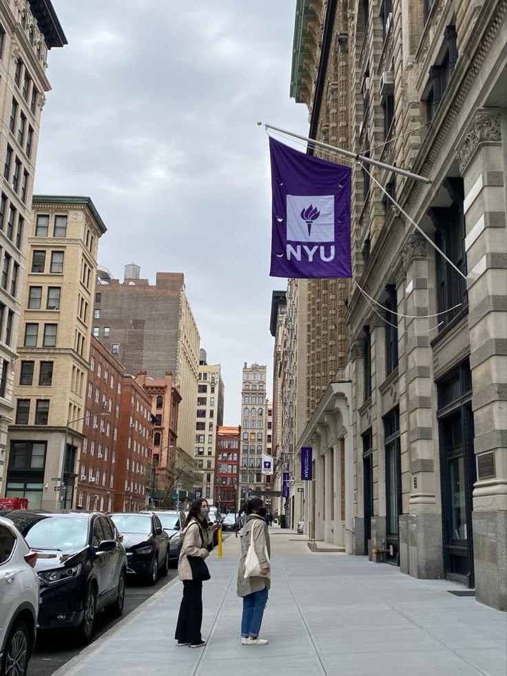 two women walking down the sidewalk in front of buildings with a purple flag hanging from it's side