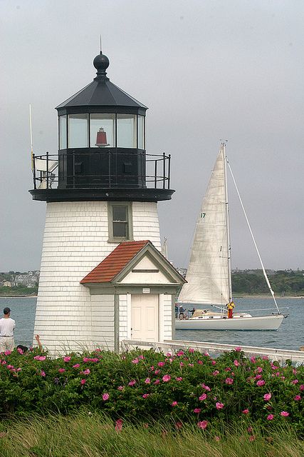 a white lighthouse with a sailboat in the water and people standing near by it