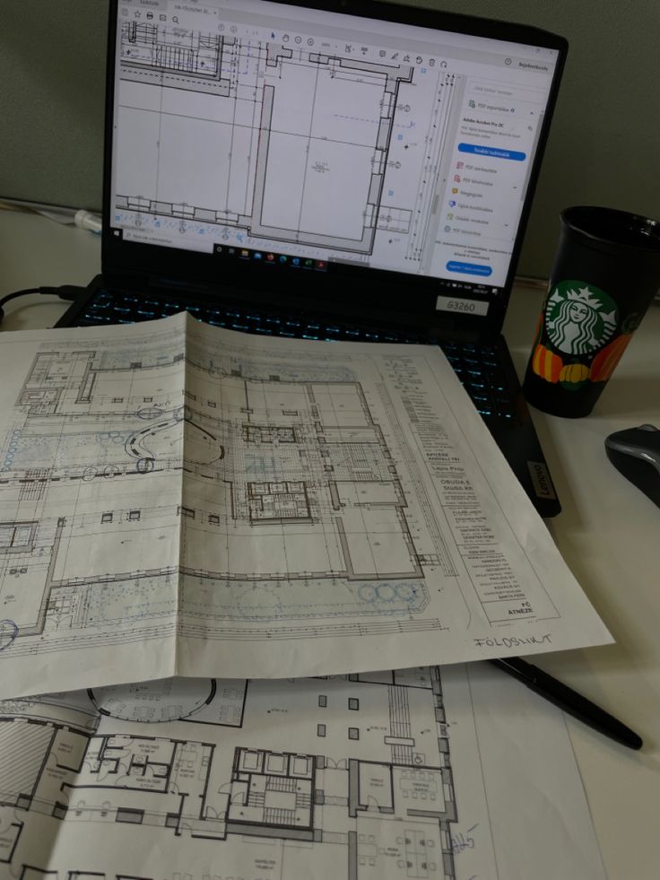 an open laptop computer sitting on top of a desk next to architectural drawings and a cup of coffee