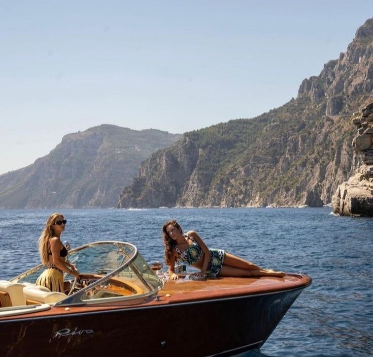 two women are sitting on the back of a boat in the water near some mountains