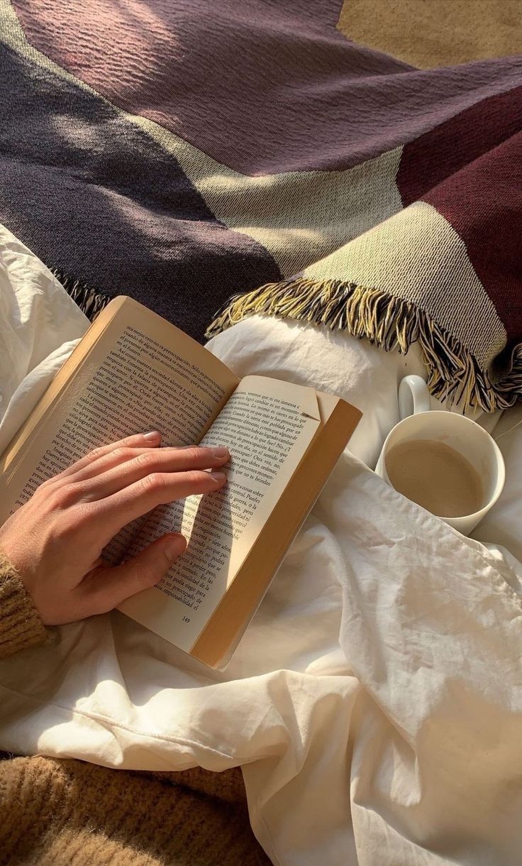 a person is reading a book and drinking coffee on a bed with blankets around them