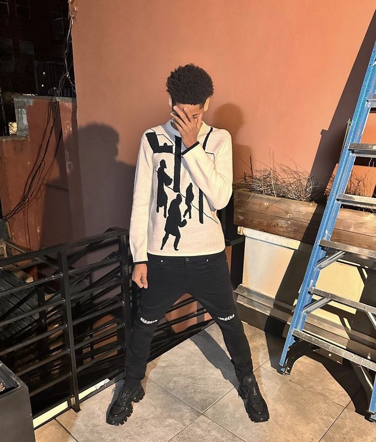 a young man standing in front of a wall with his hands to his face while wearing black pants and a white sweatshirt