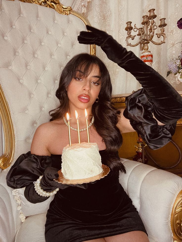 a woman sitting in a chair holding a cake with candles on it and wearing black gloves