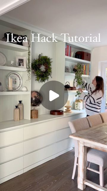 a woman standing in front of a kitchen counter with white cabinets and shelves filled with dishes