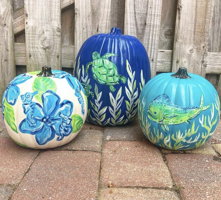 three painted pumpkins sitting next to each other on a brick floor in front of a wooden fence