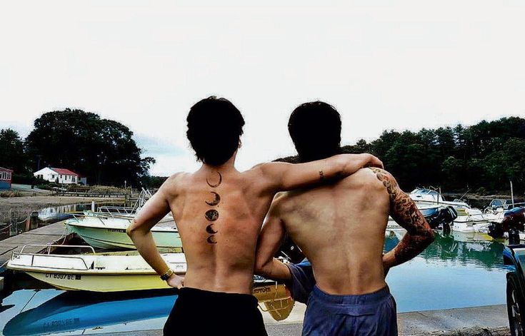 two men standing next to each other with tattoos on their backs, looking at boats in the water