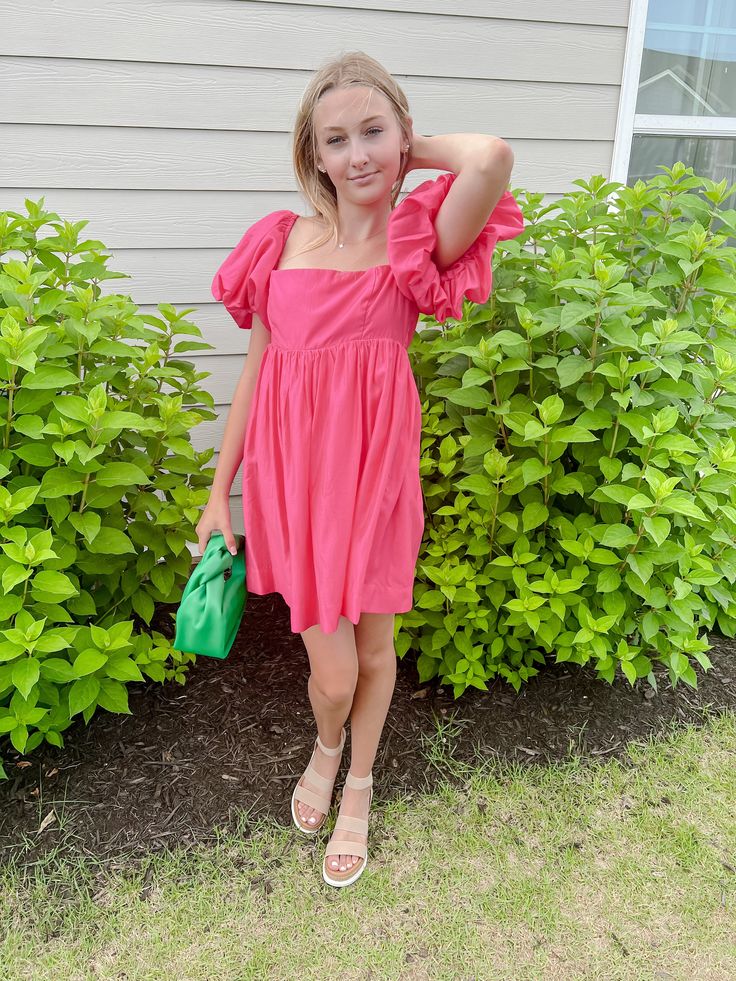 a woman standing in front of some bushes with her hand on her head and holding a green purse