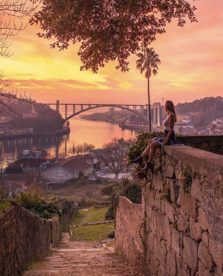 a woman sitting on top of a stone wall next to a lush green hillside under a sunset