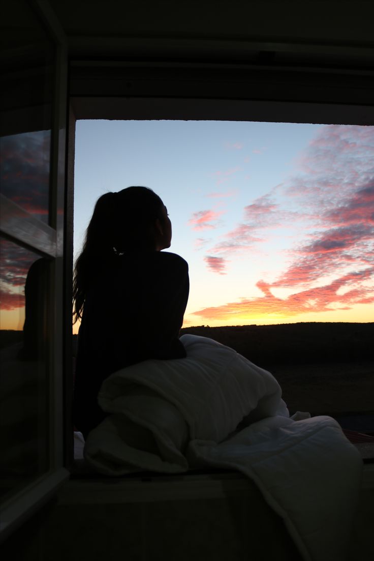 a woman sitting on top of a bed in front of a window at sunset or dawn